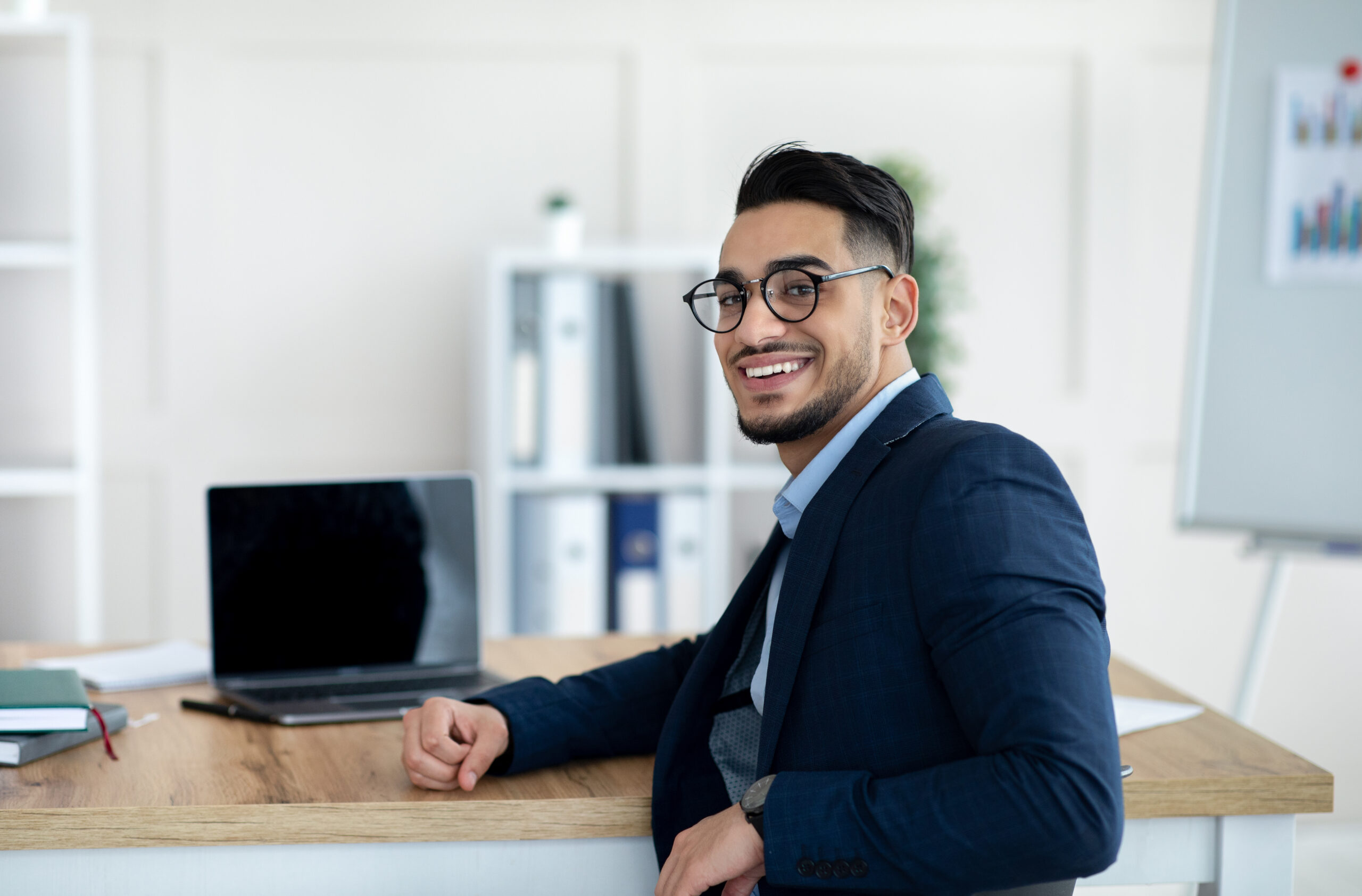 A smiling young professional in a modern office setting, sitting at his workstation with a laptop on his table.